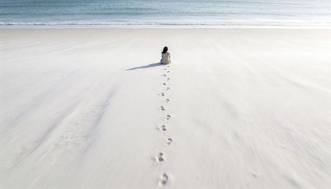 Solitary woman sitting on vast white beach leaving footprints toward calm ocean horizon