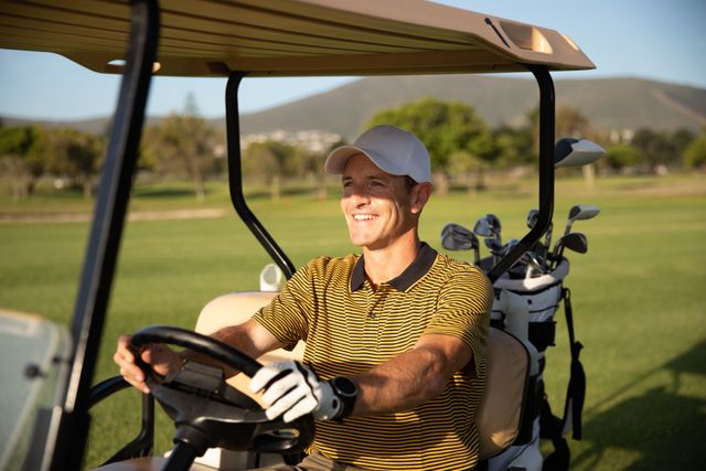 Smiling Caucasian male golfer practicing on a golf course on a sunny day wearing a cap and golf clothes, driving a golf cart. Hobby healthy lifestyle leisure.