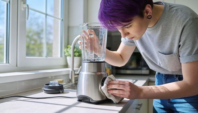 Purple-haired young adult cleaning stainless steel blender in sunlit minimalist kitchen