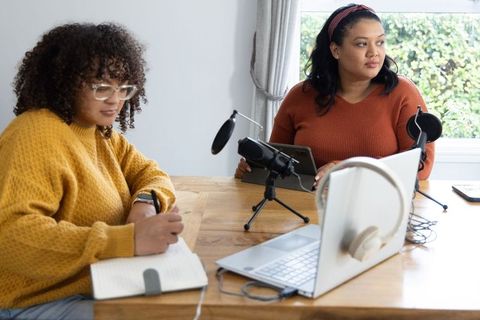 Female colleagues recording podcast and taking notes in home workspace