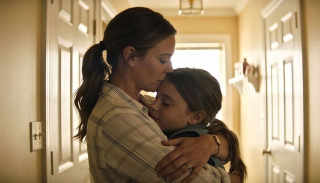 Tender mother kissing daughter's forehead while hugging in sunlit hallway, family intimacy
