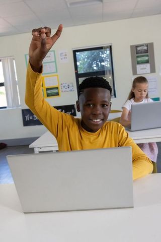 Elementary student raising hand while using laptop in bright classroom