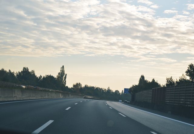 Wide-angle view of a deserted highway under a bright and clear sky with scattered clouds. Trees line both sides of the asphalt road, adding a touch of natural beauty. Ideal for themes of travel, freedom, and journeying as well as articles on transportation infrastructure.