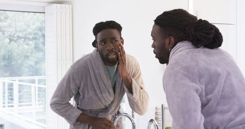 African american man inspecting cheek in bathroom mirror wearing gray bathrobe