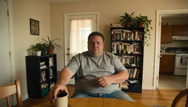 Man sitting at kitchen table reaching for mug in cozy sunlit home with bookshelves