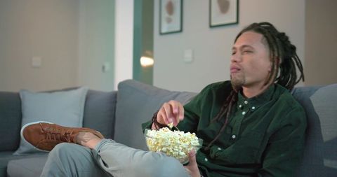 Mid-adult man reclining on modern couch holding bowl of popcorn watching tv in living room