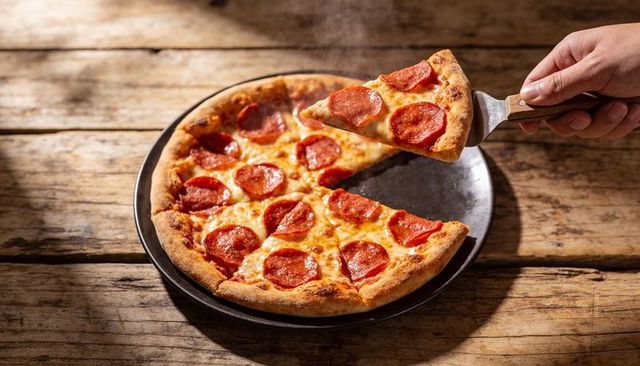 Lifting pepperoni pizza slice with spatula from steaming rustic pizza on wooden table