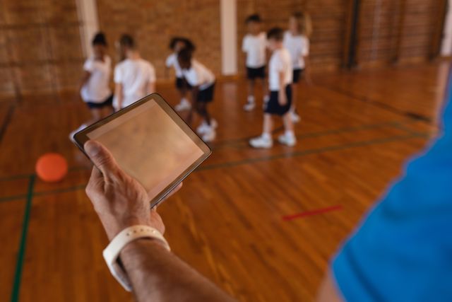 Basketball coach using digital tablet while students practice in school gym. Useful for illustrating modern coaching techniques, integration of technology in sports education, and physical education activities in schools.
