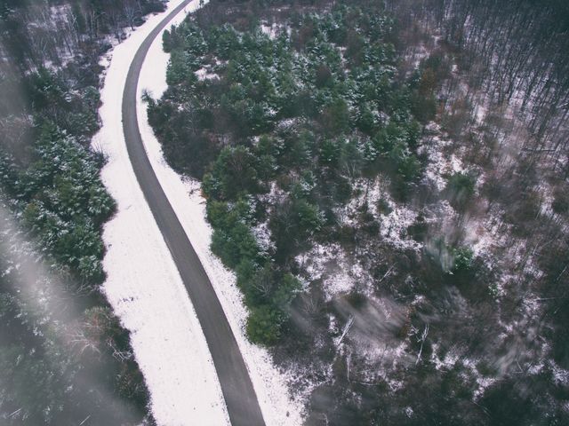 Aerial view of a snow-covered road winding through a forest during winter. Dense trees on either side with a fresh layer of snow create a serene winter landscape. Ideal for use in travel blogs, environmental articles, winter-themed advertisements, nature documentaries, and outdoor adventure promotions.