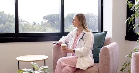 Female clinician taking coffee break in modern clinic waiting room by window, stethoscope