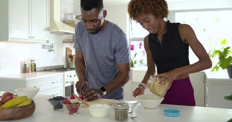 Couple preparing healthy breakfast together pouring granola slicing strawberries at kitchen island