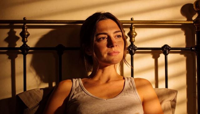 Sunlit woman sitting on bed gazing through blinds with striped shadows, warm golden light