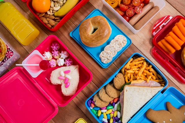 Top view of a colorful lunch box arrangement with diverse snacks and sweets on a wooden table. Ideal for illustrating meal prep, school lunches, balanced diet, and healthy eating habits for children. Useful for blog posts, educational content, meal planning guides, and advertisements for food products or lunch containers.