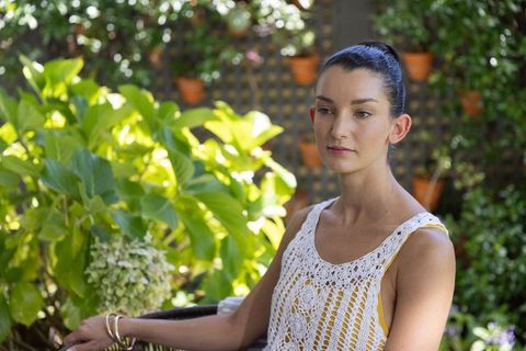 Calm woman sitting on wicker chair wearing crochet top in sunny garden with clay pots