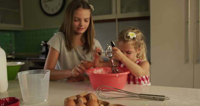 Two young sisters baking together in a cozy kitchen. One girl assisting younger sibling with mixing ingredients in a bowl. Eggs and kitchen utensils visible on counter. Perfect for family, cooking, and childhood themed projects, advertisements, or publications.