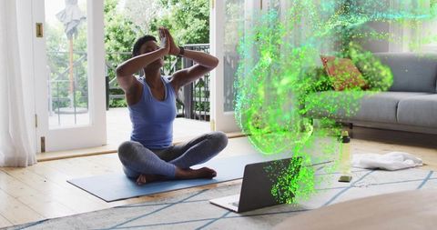 Woman practicing guided meditation on yoga mat with laptop and green digital aura, sunlit balcony