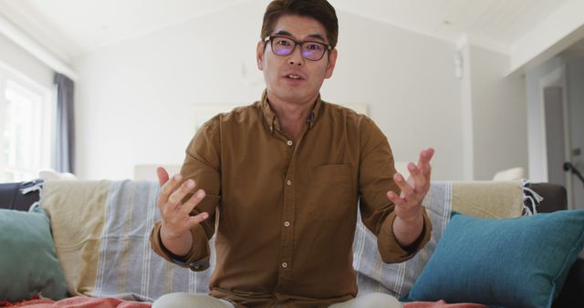 Asian man sitting on couch, talking excitedly while making video call from home. Useful for visualizing remote communication, online meetings, or modern lifestyle contexts. Suitable for technology, communication services, or home comfort themes.