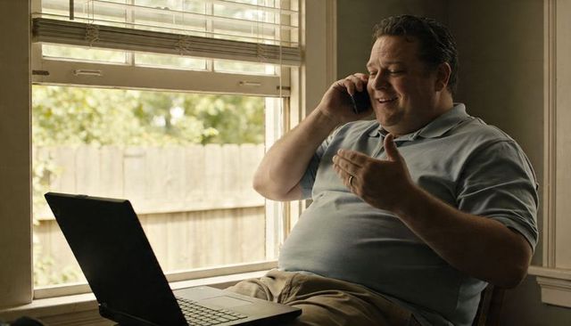 Smiling man talking on phone while using laptop by sunlit window at home