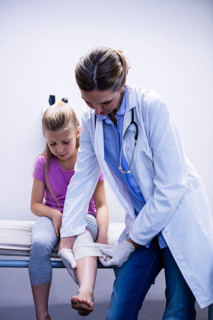 Doctor bandaging young girl's injured leg in hospital. Ideal for use in healthcare, medical care, pediatric care, and emergency care contexts. Can be used in articles, brochures, and websites related to medical treatment, first aid, and child healthcare.