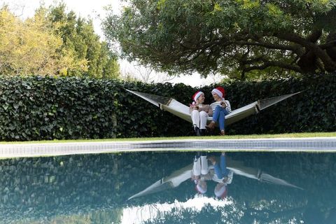 Diverse women wearing santa hats relaxing on hammock beside backyard pool, holiday rest