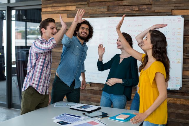 Group of young business executives celebrating success by giving high fives in a modern office. They are standing around a table with documents and a whiteboard filled with notes in the background. Perfect for illustrating teamwork, collaboration, and a positive work environment in corporate or startup settings.