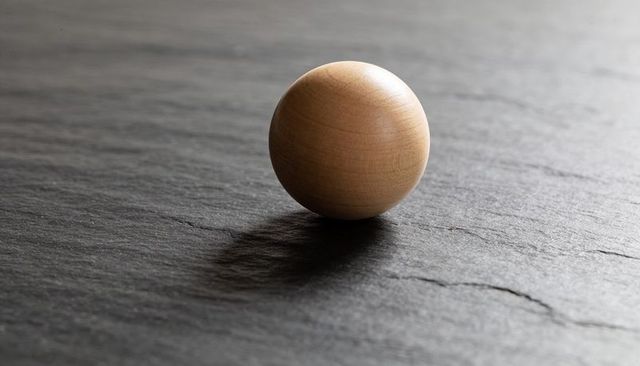 Minimal wooden sphere on dark slate surface catching studio highlight and soft shadow