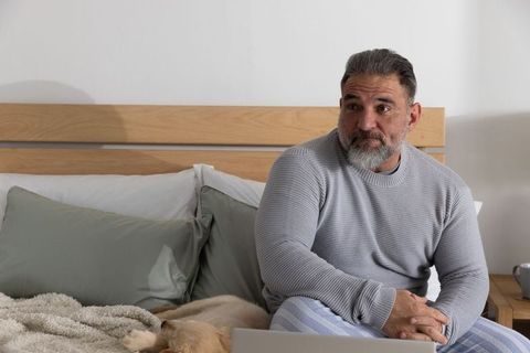 Mature bearded man sitting on bed using laptop while dog resting, cozy morning routine