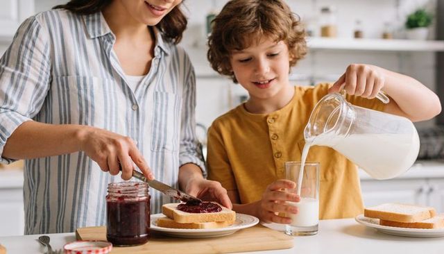 Mother spreading jam while son pouring milk bright kitchen morning family bonding