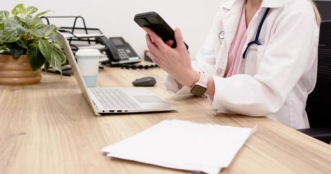 Female physician checking smartphone at desk with laptop and paperwork in modern clinic