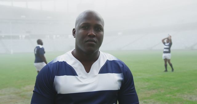 Portrait of bald african american male rugby player in strip on stadium pitch, players in background. Rugby, sport, sportsman, confidence and competition, unaltered.