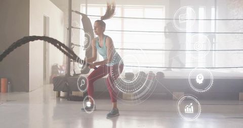 Female athlete swinging battle ropes in boxing ring studio backlit fitness training