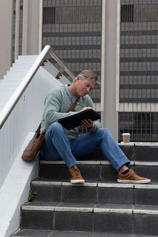 Middle-aged man sketching on city steps with coffee cup and leather satchel