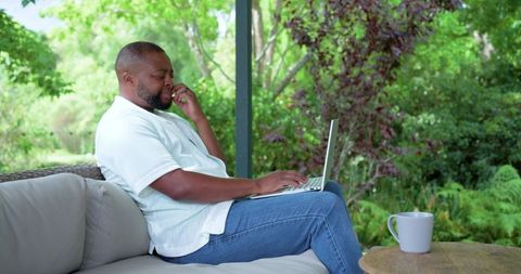 African man working remotely on laptop and talking on phone on garden porch with coffee