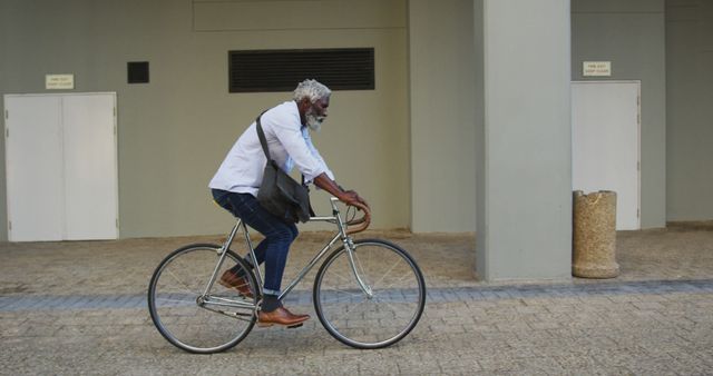 Elderly man is riding a bicycle on a city street, carrying a shoulder bag. He is wearing denim pants, a white shirt, and brown shoes. This image can be used for articles on healthy lifestyles, urban commuting, sustainable transportation, and senior active lifestyles.