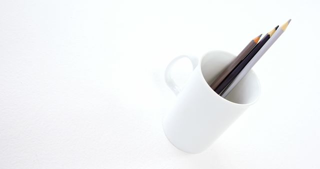A white mug holding three pencils placed on a white background. Ideal for illustrating concepts of minimalism, organization, simplicity, or creative workspaces. Suitable for use in design projects, office decor inspiration, or stationery advertisements.