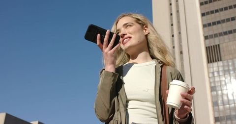 Young woman speaking into smartphone while holding takeaway coffee near office tower