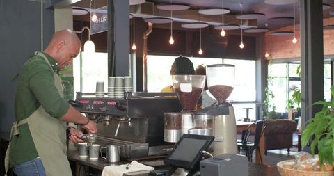 Barista preparing espresso behind counter with grinders, pos terminal and cozy cafe interior