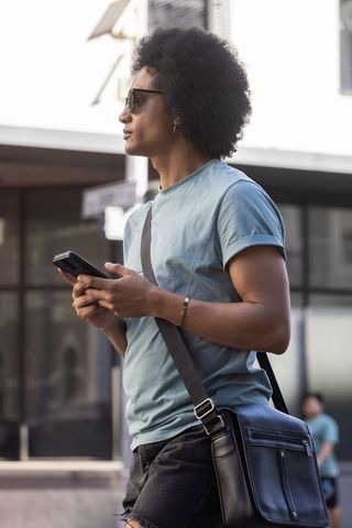 African american man standing on city sidewalk holding smartphone wearing crossbody bag