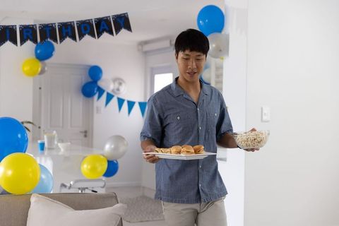 Asian man holding plate of sliders and bowl of popcorn at blue yellow birthday party