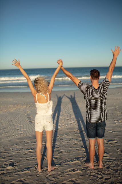 This image captures a couple standing on the beach with their arms raised, symbolizing joy and freedom. Ideal for use in travel brochures, romantic getaway promotions, lifestyle blogs, and advertisements focusing on summer vacations, relationships, and outdoor activities.
