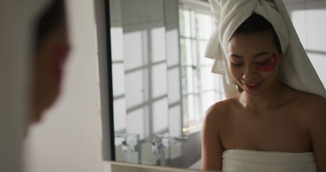 Vertical image of smiling biracial woman with towel on hair looking in mirror in bathroom. Health and beauty, leisure time, domestic life and lifestyle concept.