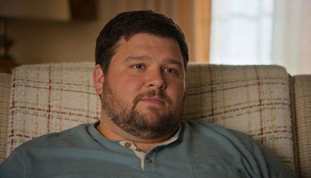 Bearded man sitting on plaid couch gazing through sheer curtain window contemplative moment