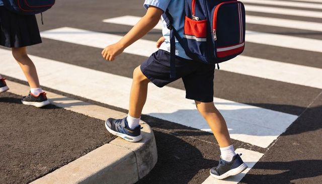 Elementary students crossing street wearing uniforms and backpacks at crosswalk sunlit curb