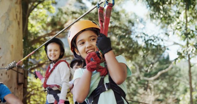 Children experiencing the thrill of a zipline high up in the trees, wearing comprehensive safety gear including helmets and harnesses. They look excited and enjoy the exhilarating outdoor activity. Ideal for promoting family outings, adventure sports, kid-friendly activities, summer camps, and safe recreational programs.