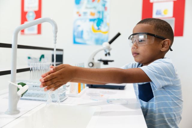 African american elementary schoolboy wearing protective eyewear washing hands in laboratory. unaltered, education, learning, laboratory, stem, scientific experiment, protection and school concept.
