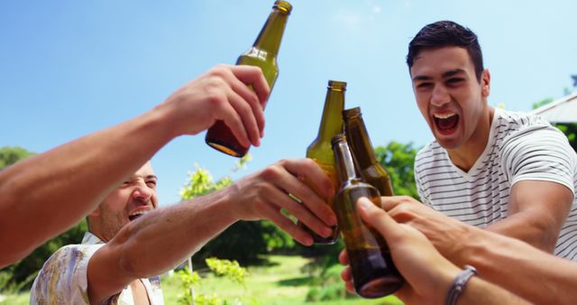 Group of male friends enjoying sunny day, raising beer bottles in toast, smiling and laughing outdoors. Ideal for content related to celebrations, parties, friendship, summer activities, outdoor gatherings, or advertisements promoting drinks and beverages.