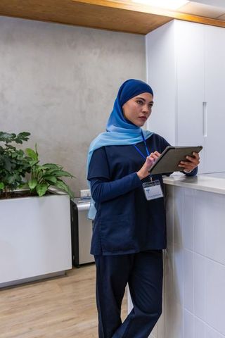 Muslim nurse wearing hijab using tablet at clinic reception leaning on counter healthcare