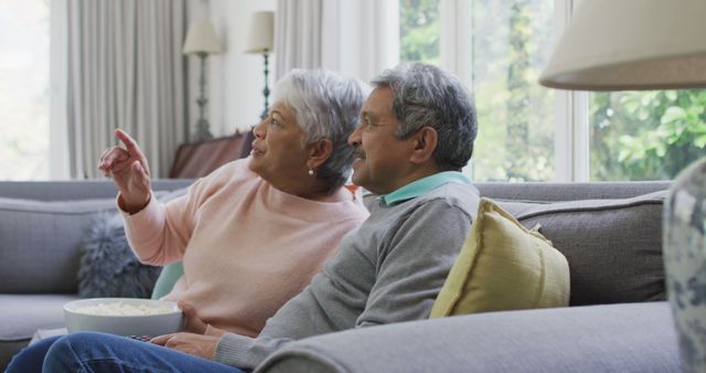 Elderly man and woman sitting and relaxing together on couch in living room, both watching television. They're enjoying snacks and conversing. This can be ideal for advertisements about family life, retirement communities, and healthy aging. It suggests happiness, companionship, and comfort at home.