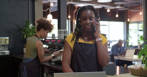 African american barista working at cafe counter using laptop and talking on phone