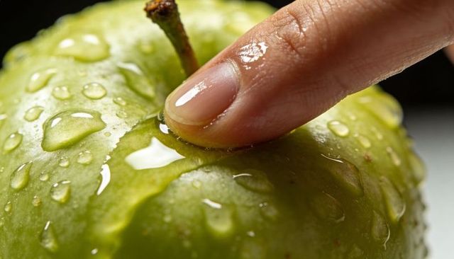 Pressing fingertip on wet green apple macro showing glossy droplets and tactile texture